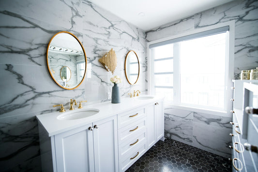 Modern bathroom with marble wall tiles, a white double-sink vanity with gold faucets, round mirrors, and hexagonal black floor tiles, featuring elegant and luxurious design elements.